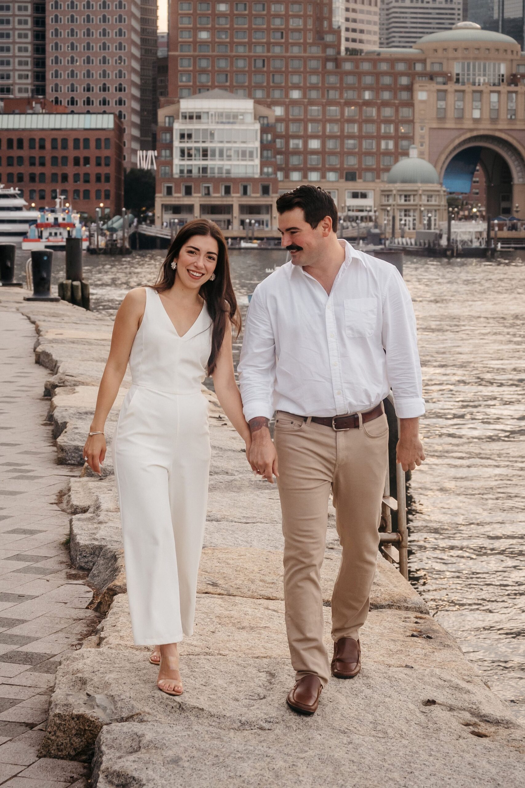 Engaged couple walking hand in hand at Fan Pier Park Boston engagement session, city skyline and waterfront backdrop at sunset