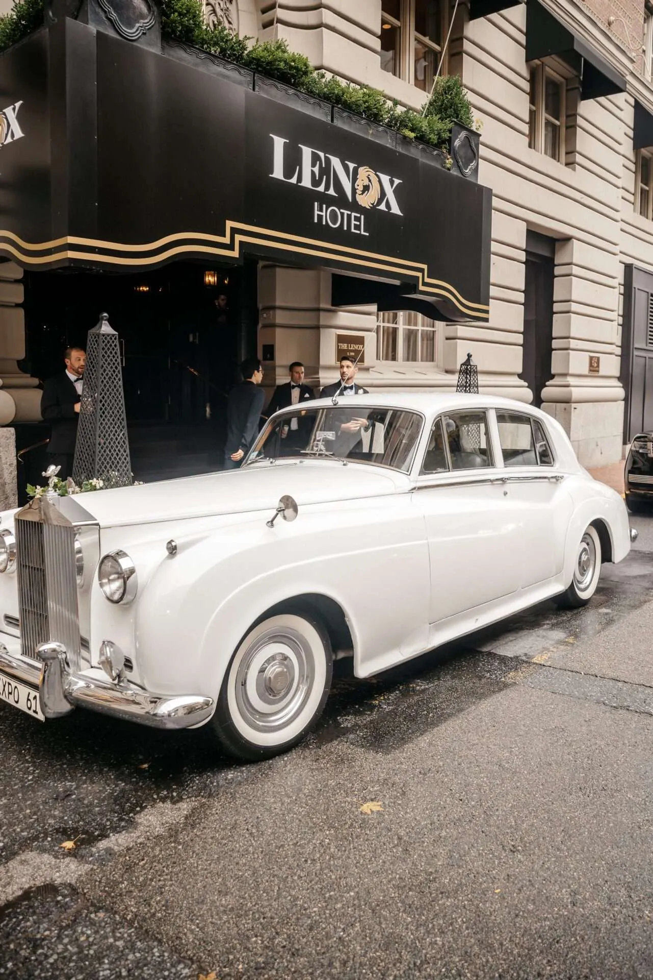A white vintage car is parked in front of the lenox hotel while a bride and groom are getting married in massachusetts