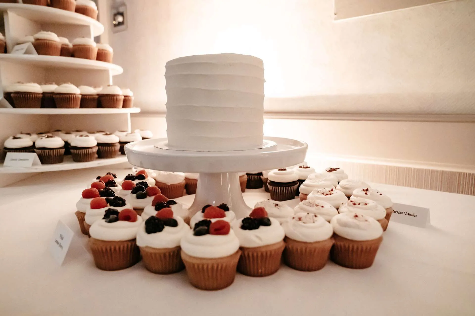 A white wedding cake sits on a cake stand surrounded by cupcakes in burlingon wedding locations