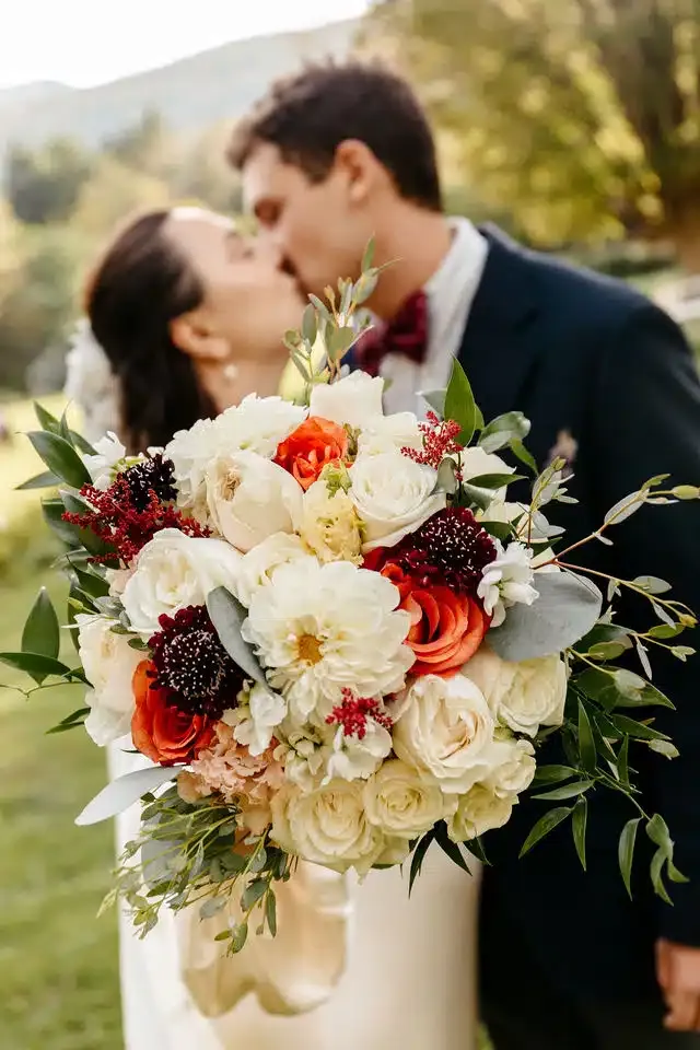 A close-up shot of a lush wedding bouquet featuring white roses, dahlias, and deep red and orange autumn flowers, with a bride and groom sharing a blurred kiss in the background