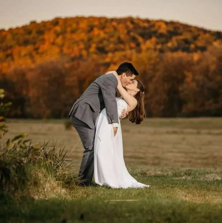 A groom in a grey suit dips and kisses his bride in an open field against a scenic background of vibrant New England autumn foliage trees.