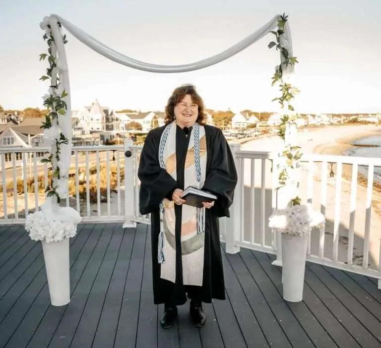A female wedding officiant stands smiling at a decorated outdoor altar on a white deck overlooking the ocean beach at a waterfront Falmouth venue.