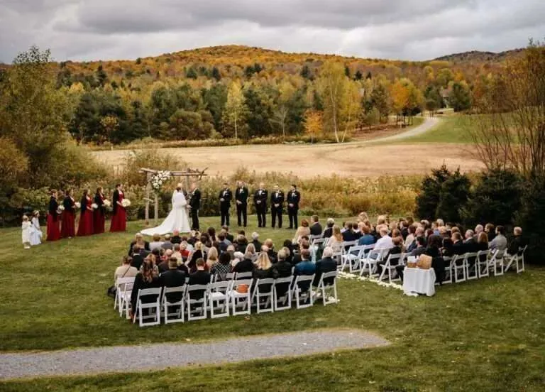 A wide, scenic landscape view of an outdoor wedding ceremony setup tucked into a vast field surrounded by rolling hills covered in peak New England fall foliage.
