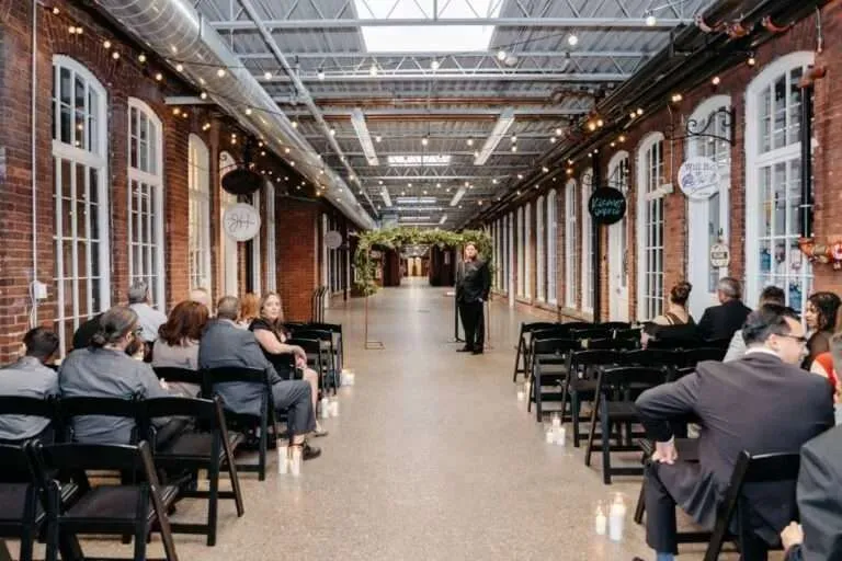 An elegant indoor wedding ceremony setup inside a historic brick mill venue featuring long rows of black chairs, hanging string lights, and a greenery arch at the altar.