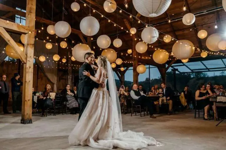 A bride and groom share their romantic first dance inside a rustic barn venue illuminated by glowing white hanging paper lanterns and market string lights.