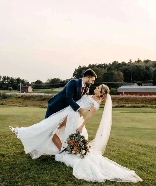 A groom dips his bride lovingly on a wide green golf course lawn during a scenic New England wedding celebration at sunset.