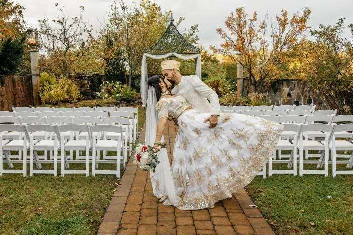A groom in elegant white and gold wedding attire dips and kisses his bride in a stunning white and gold lehenga on a brick walkway lined with white ceremony chairs in an outdoor garden venue.