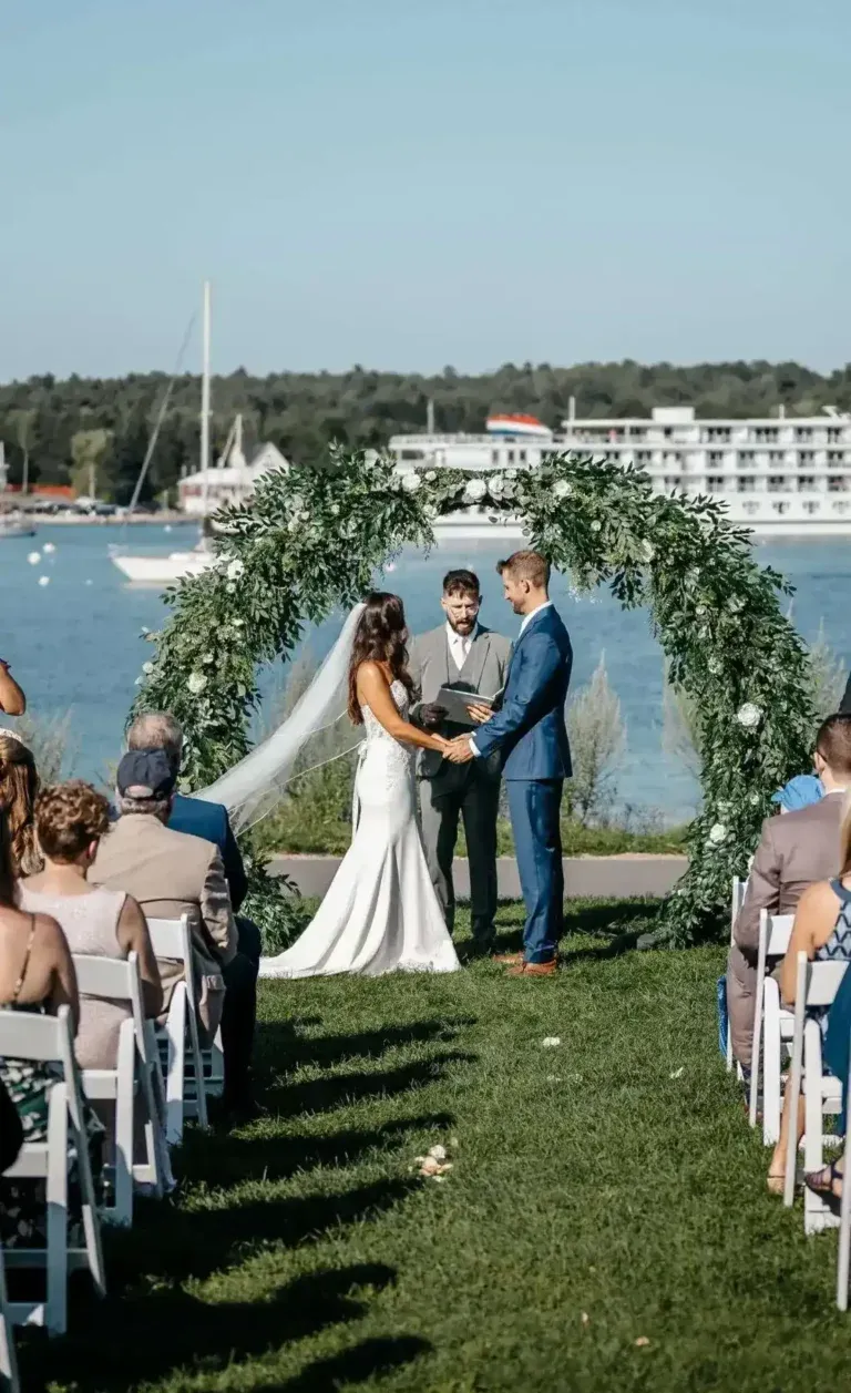 A bride and groom hold hands at the altar under a lush circular greenery arch during an outdoor waterfront wedding ceremony overlooking a harbor with boats.