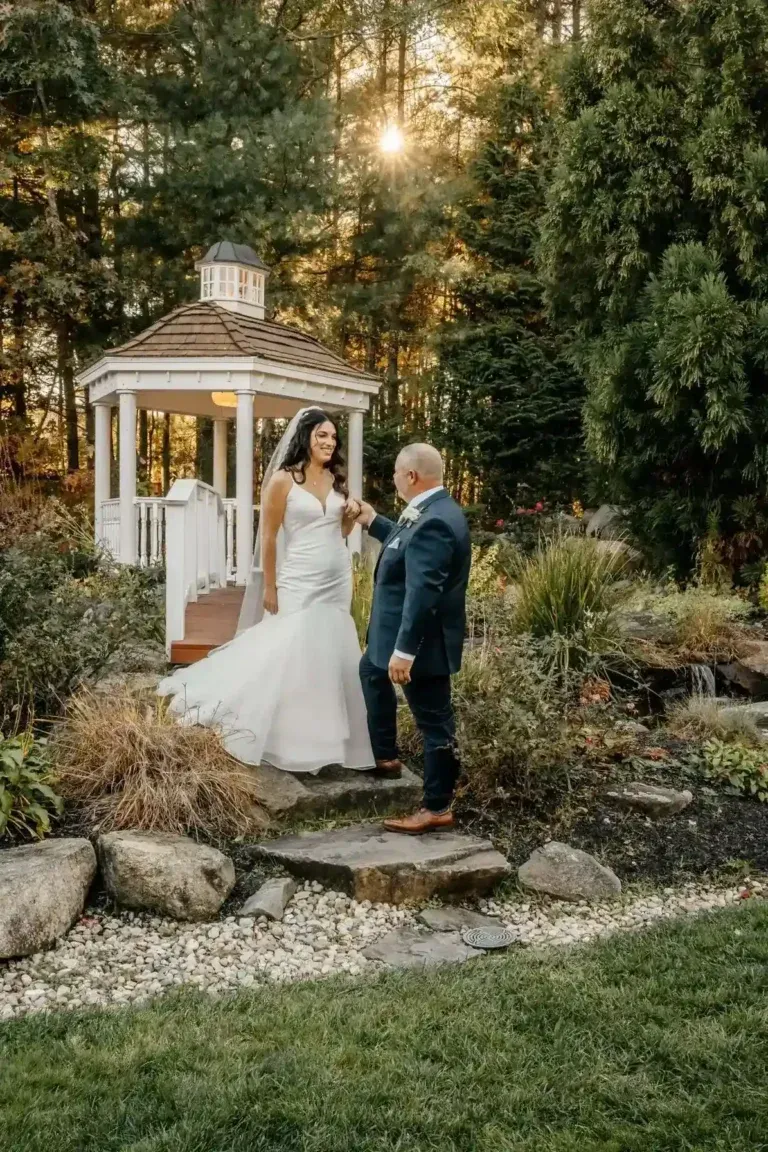 A groom in a navy suit holds his bride's hand as they pose on a stone path next to a white wooden garden gazebo with golden sunlight filtering through the trees.