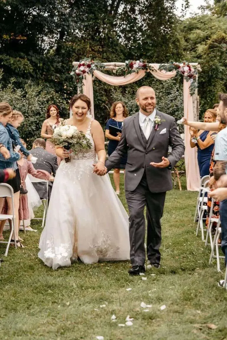 A newly married bride and groom smile joyfully while walking down the aisle at an outdoor New England wedding venue during their ceremony recessional as guests toss petals.
