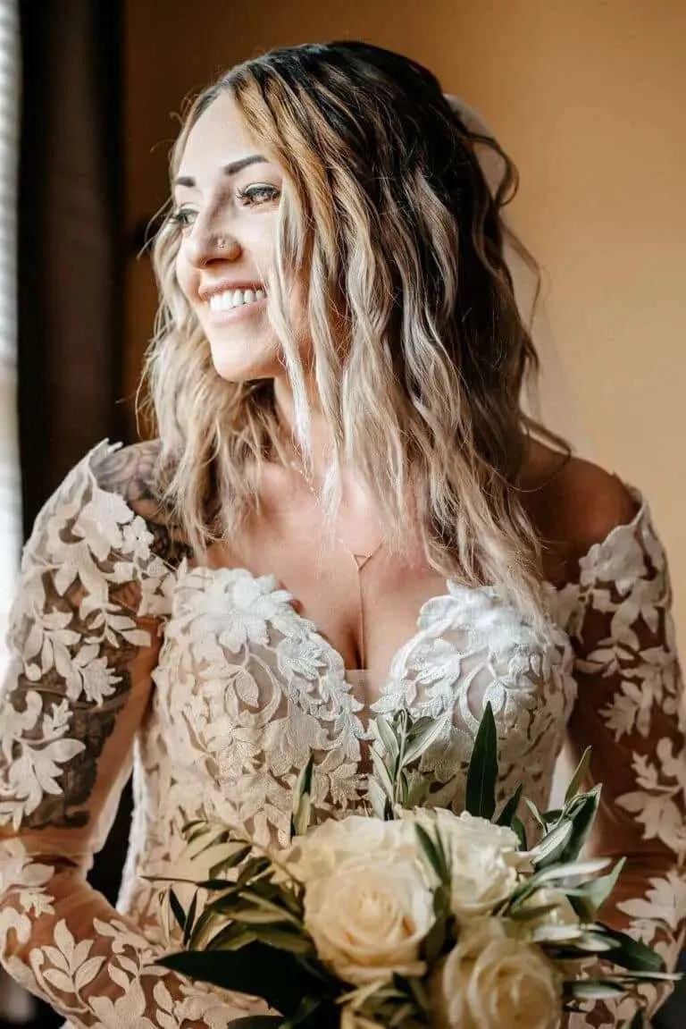 A beautiful close-up bridal portrait of a smiling bride with a tattoo sleeve wearing an elegant long-sleeve lace wedding gown and holding a white rose bouquet.