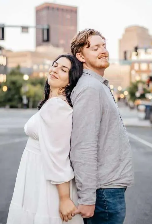 A romantic portrait of a bride in a white dress and a groom in a grey button-down shirt standing back-to-back with their eyes closed and smiling in a city center street during a New England urban wedding session at golden hour.