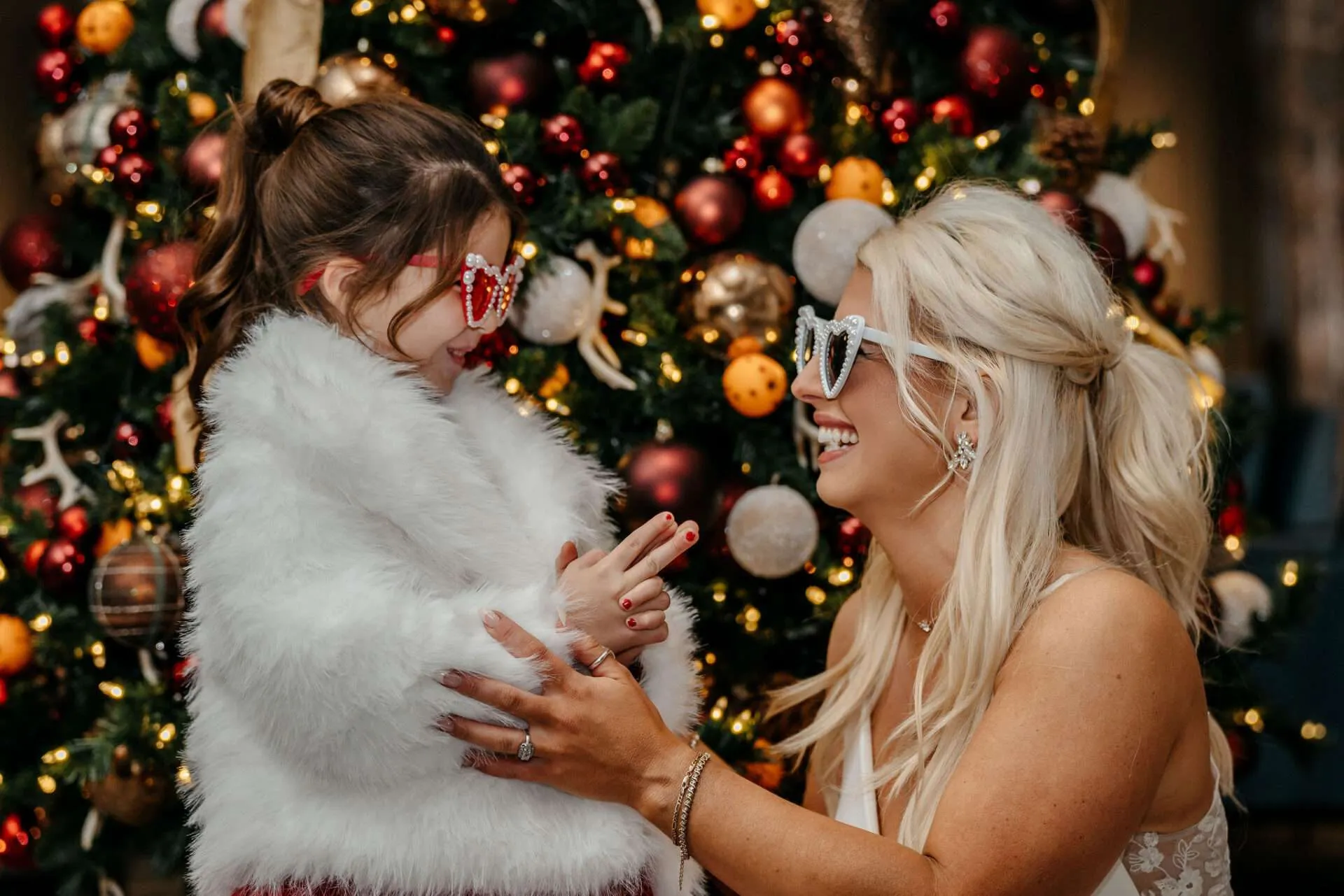 Bride and her daughter in heart-shaped sunglasses laughing by the Christmas tree