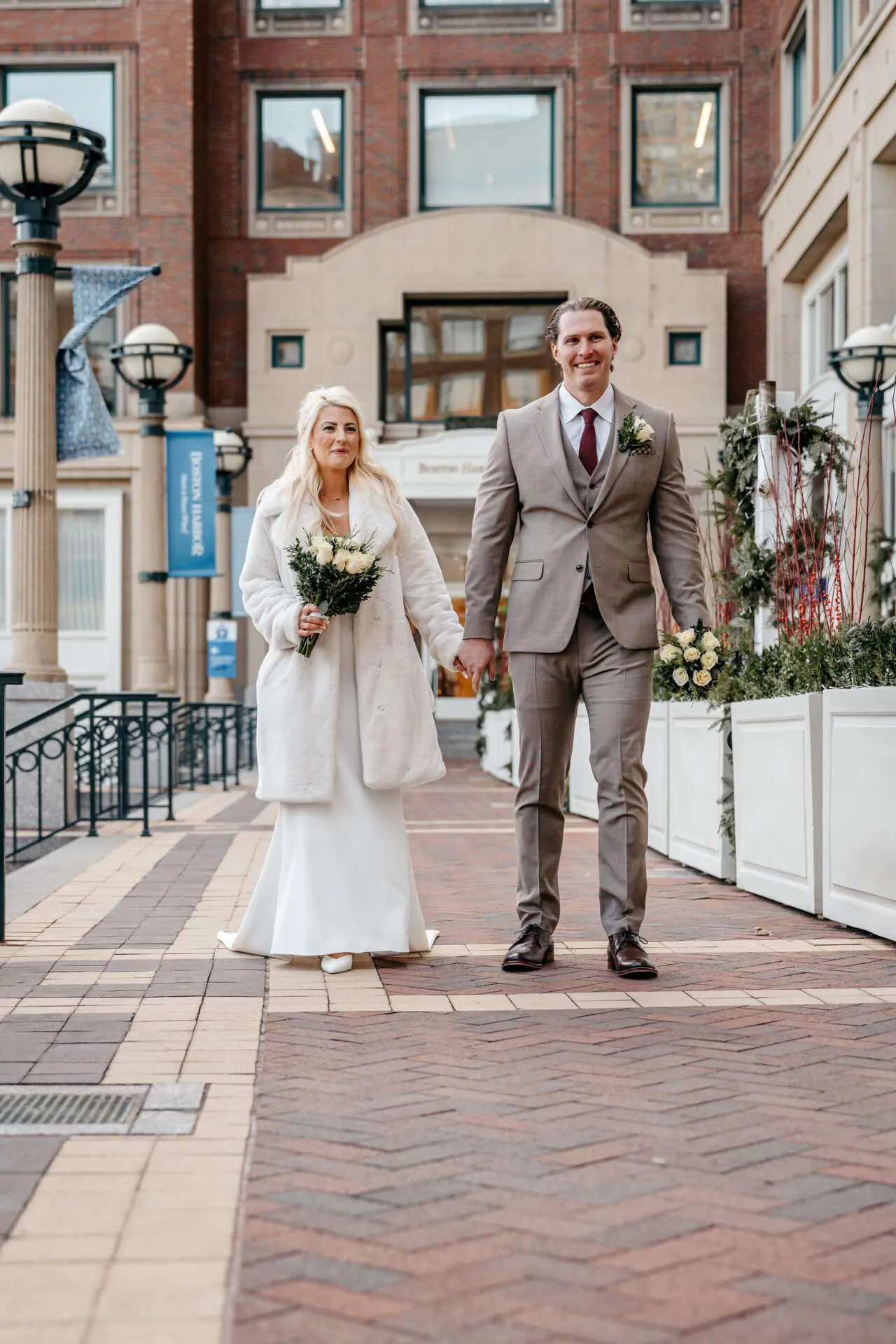 A bride and groom smile while carrying white bouquets and walk down the sidewalk to Boston city hall