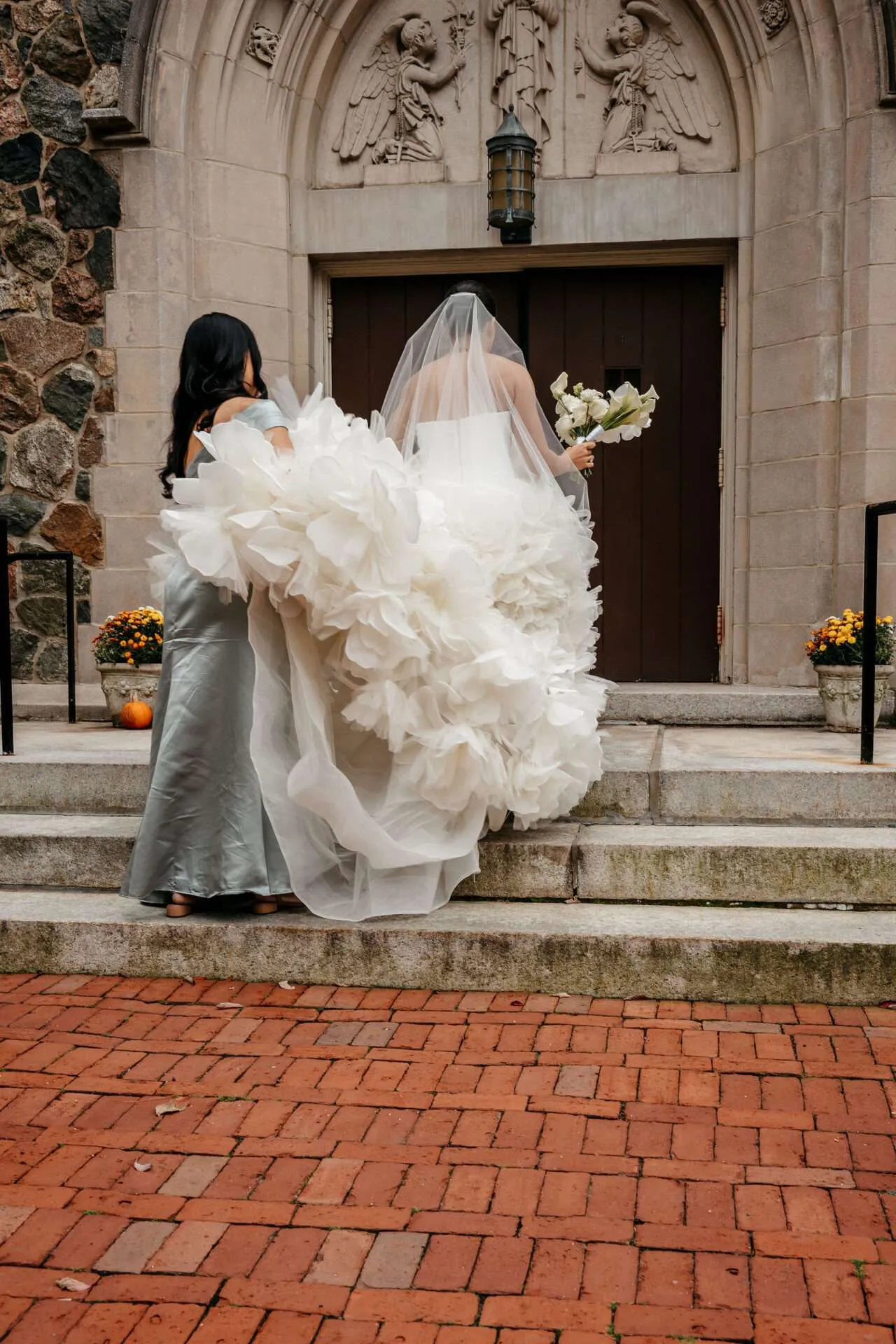A bride holding a white bouquet recieves help from her bridesmaid while climbing stairs to small wedding venues for 30 guests