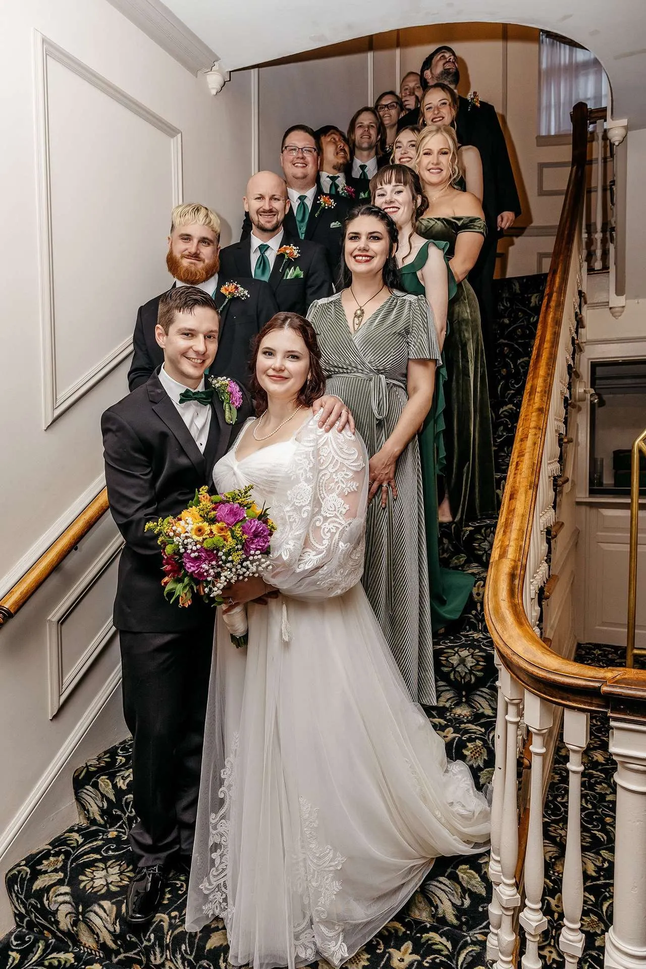 A wedding party with bridesmaids in green dresses and groomsman in black tuxedos and green ties stand on a staircase for photos at boston hotels wedding venues