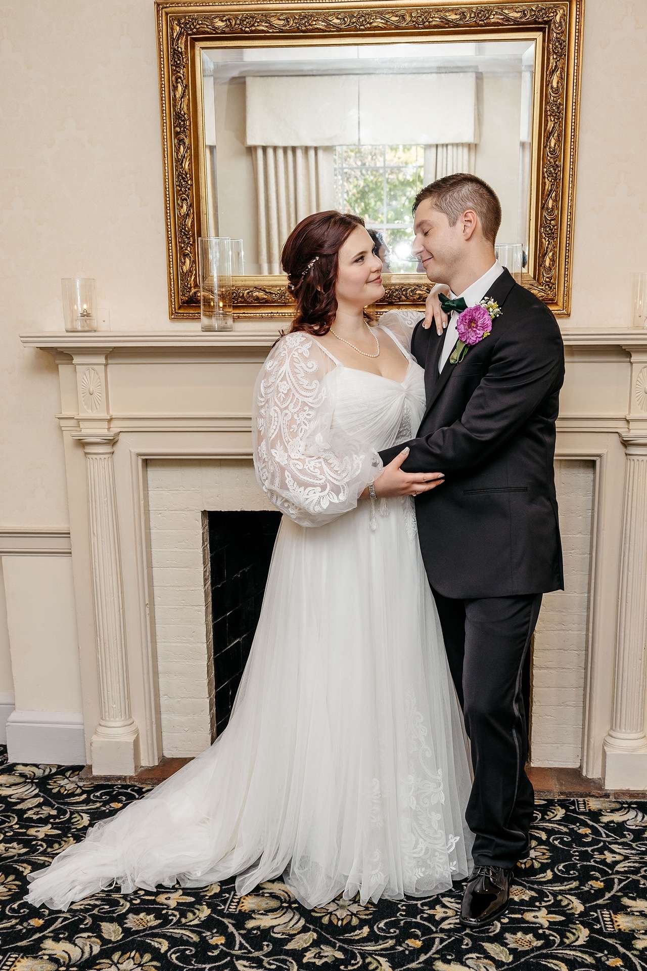 A bride and groom smile and look at each other as they pose in front of a fireplace at one of many boston hotel wedding venues.