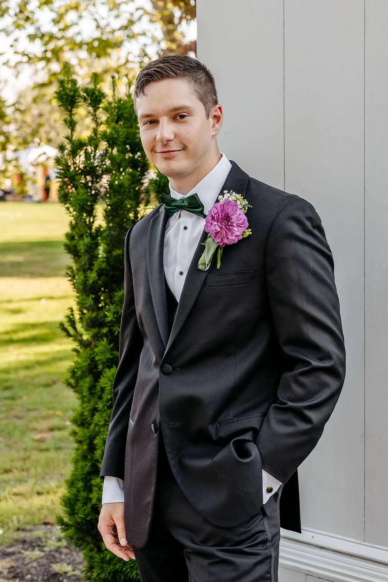 A groom wearing a black tuxedo and a purple flower stands outside posing for pictures at a boston hotel wedding venue.
