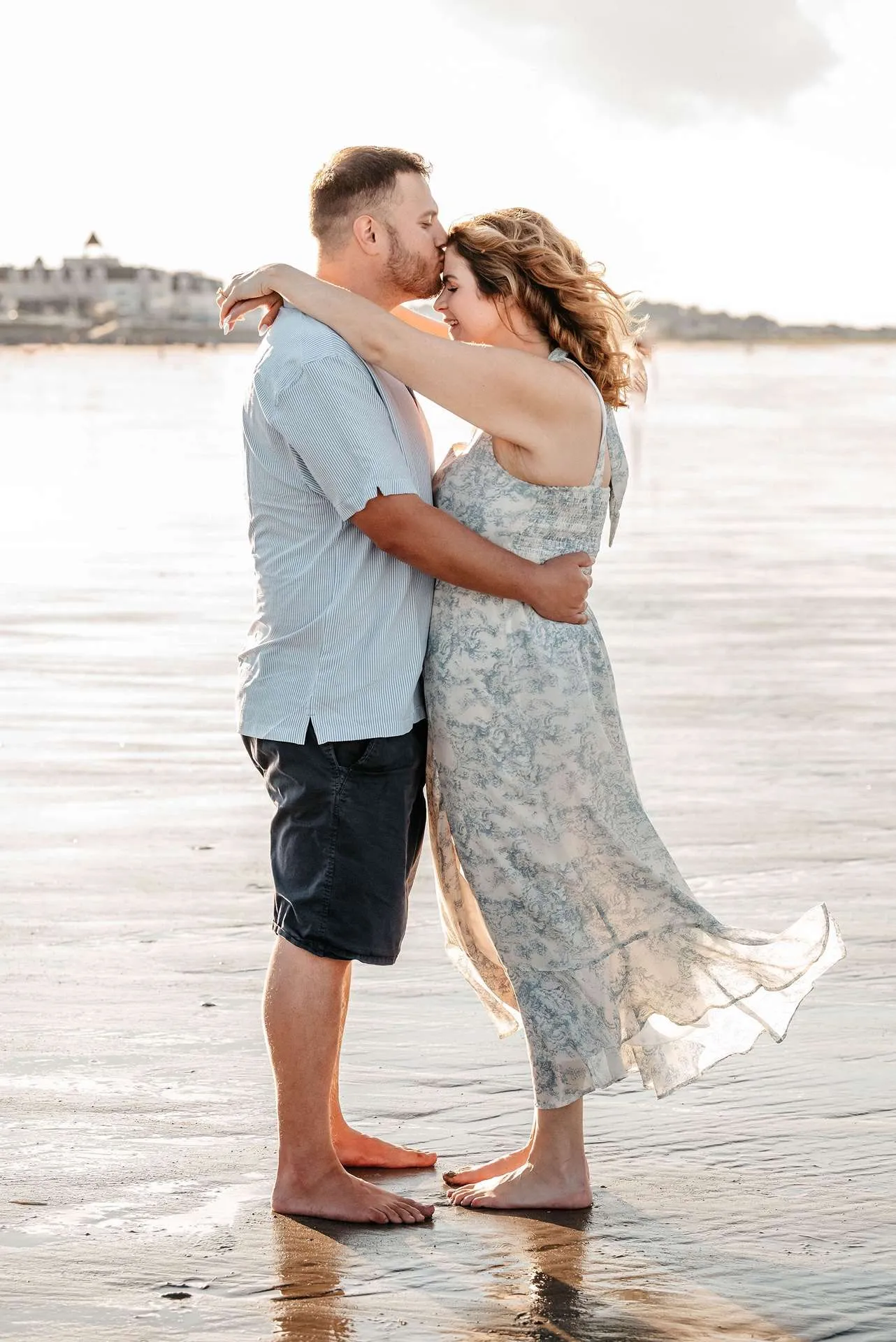 A man kisses his fiance's forehead while they embrace on the shore of a beach after deciding on some first date ideas in boston.