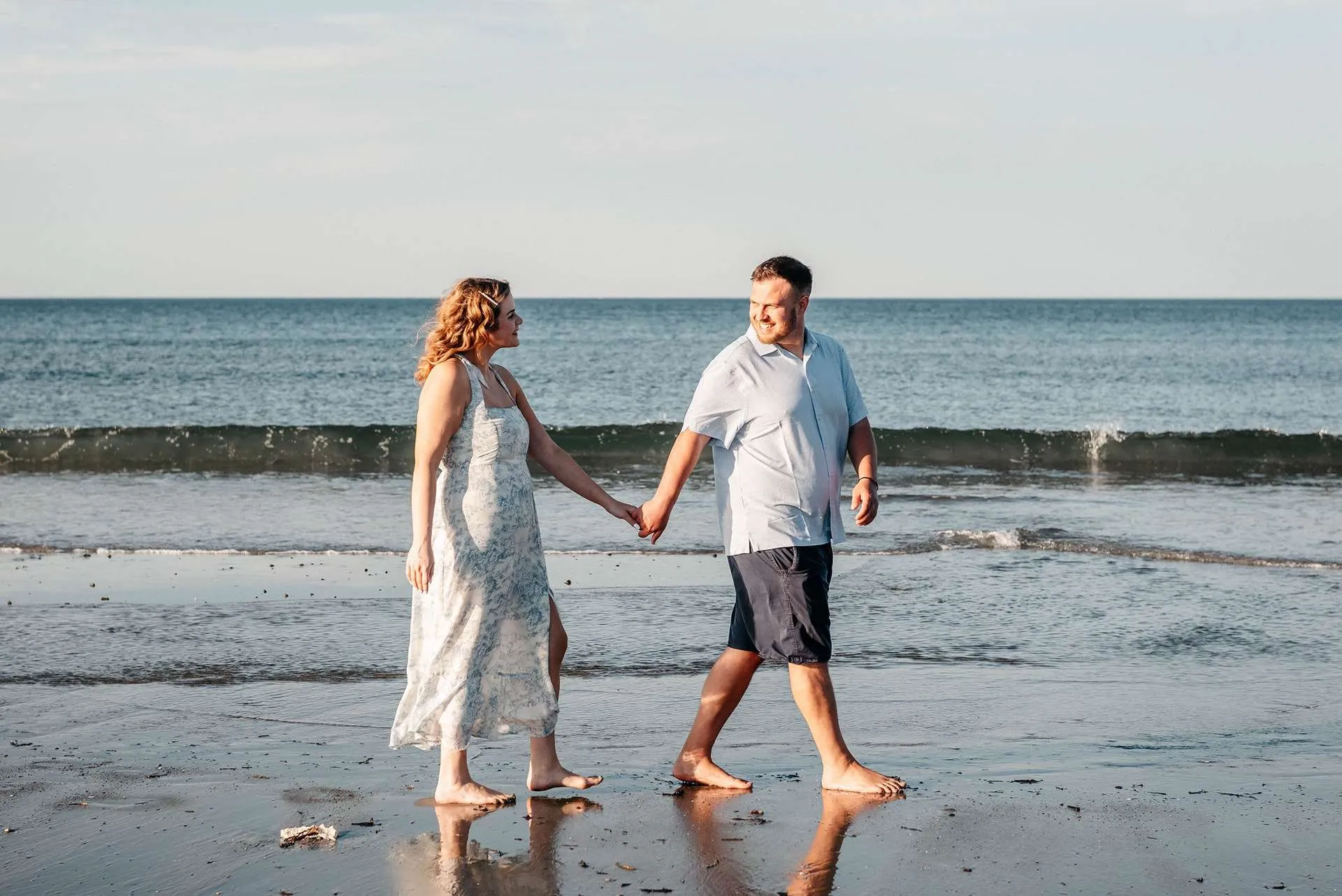 A couple in light blue outfits walk down the beach holding hands talking about date ideas in boston.