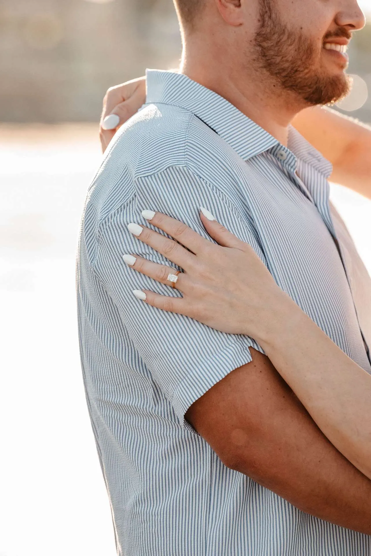 A couple stands on the beach with their engagement ring displayed on her fiance's shoulder after talking about fun date ideas in boston.