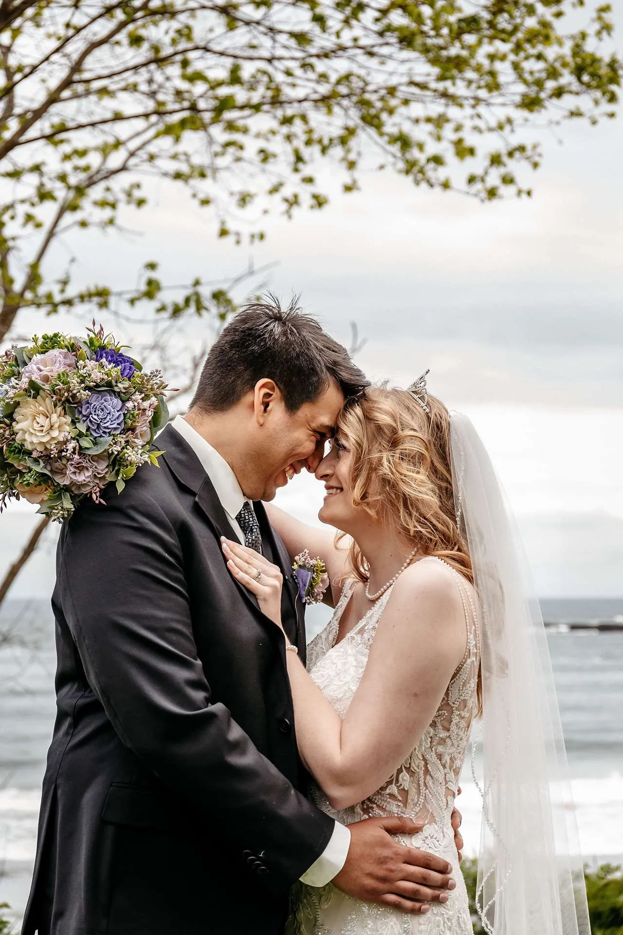 A bride and groom hold each other and smile while holding a bouquet of purple and pink flowers as a professional photographer in boston takes photos
