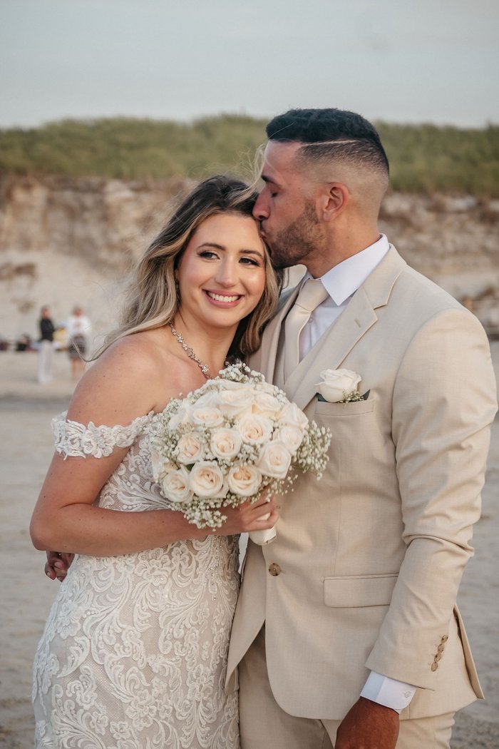 Groom kissing bride's temple on the beach at sunset – Iesha & Emanuel
