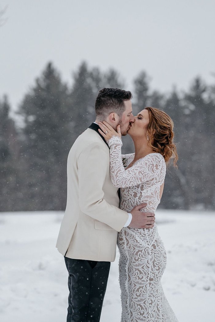 Bride and groom kissing in falling snow with pine trees behind – Seanna & Ryan winter wedding