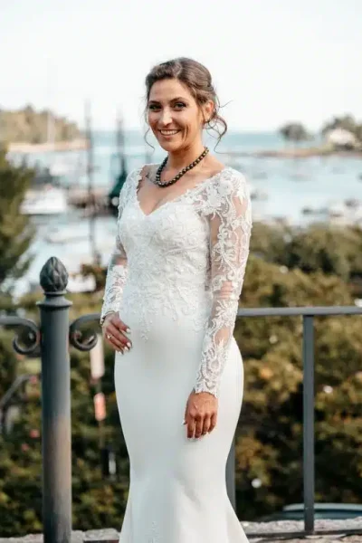 A smiling bride in a long-sleeved lace gown poses for a photo at the Camden Amphitheater in Camden, Maine.
